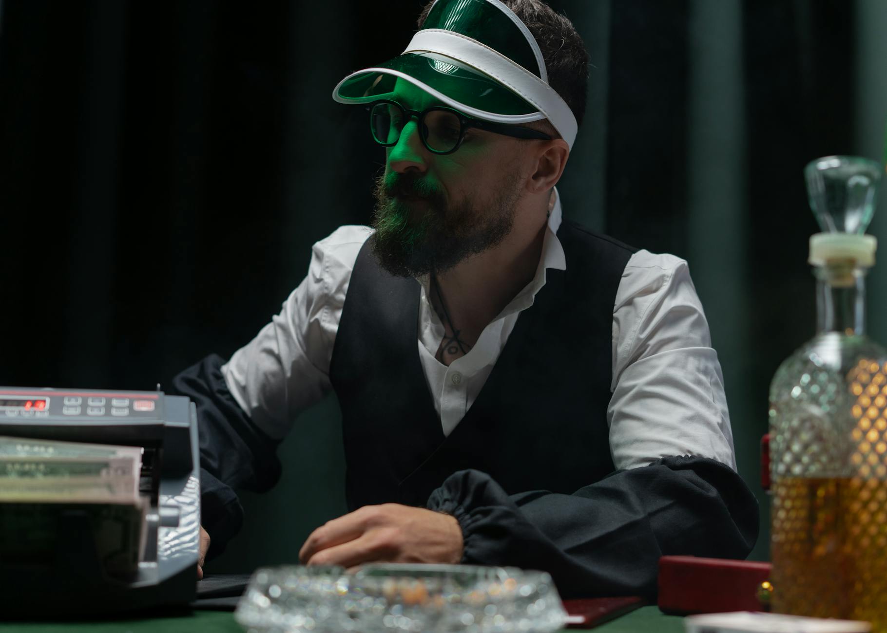 Bearded croupier in casino setting wearing visor and counting cash.