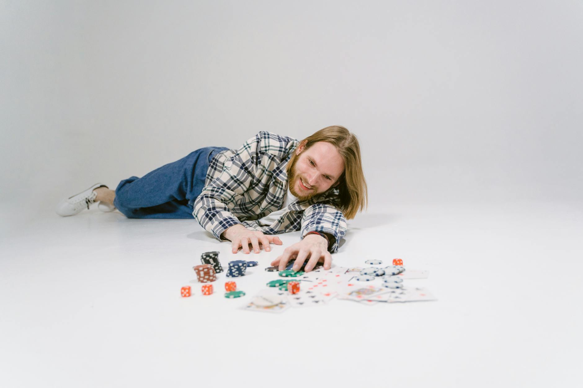 Smiling man with long hair and plaid shirt engaging with casino chips and cards on a white floor.